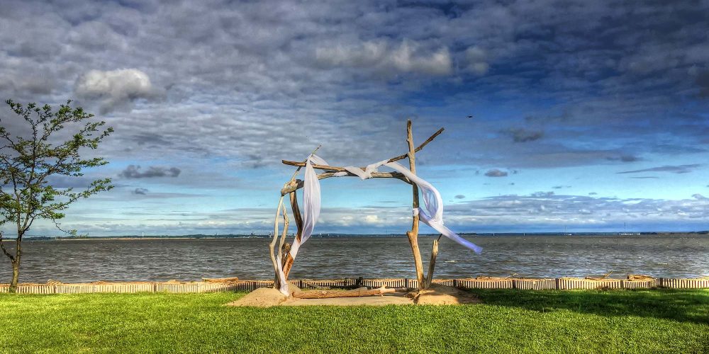 Outdoor wedding alter made from driftwood with flowing cloth blowing in the wind. It is in front of the water with a bright but cloudy sky.