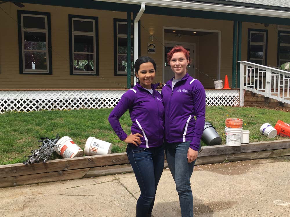 2 girls in purple jumpsuit jackets standing infront of building with buckets on grass
