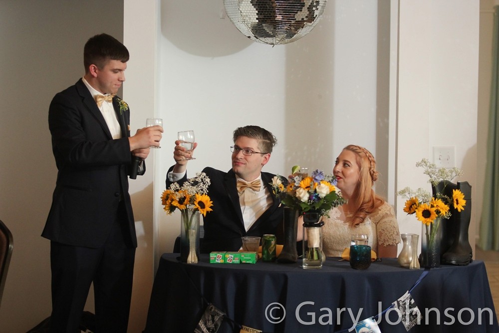 Two men in tuxes raising glases to each other next to bride